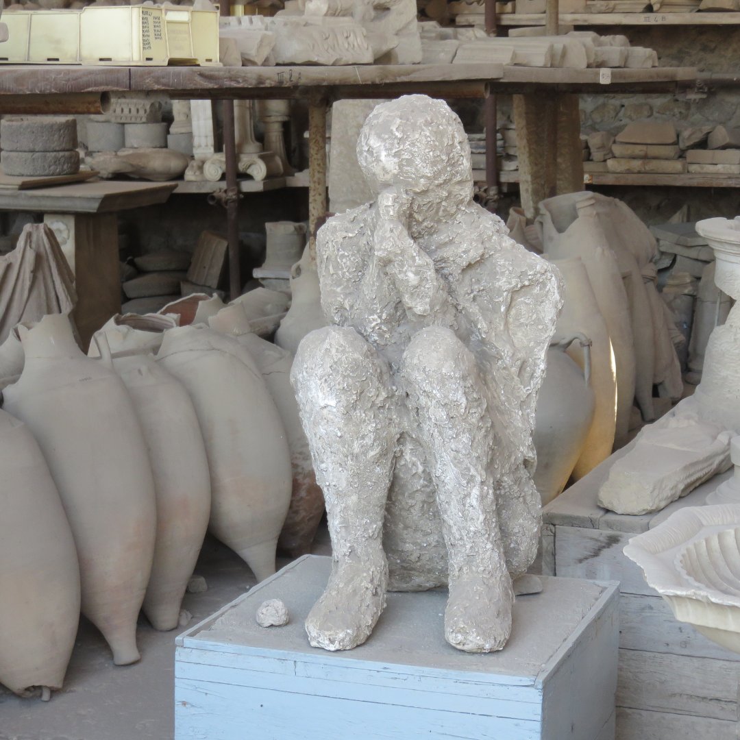 Calco in gesso di una vittima di Pompei seduta, esposto tra le anfore in un deposito